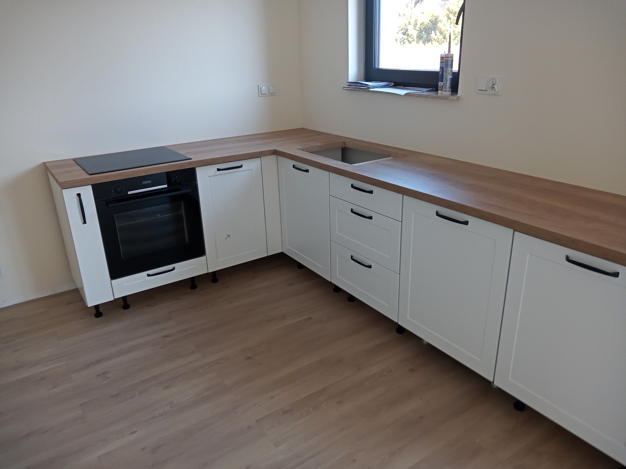 Ustawiona w rogu pomieszczenia biała, zabudowana kuchnia z drewnianym blatem, czarnymi uchwytami i wbudowaną płytą grzewczą, widoczna z boku, z oknem w tle.