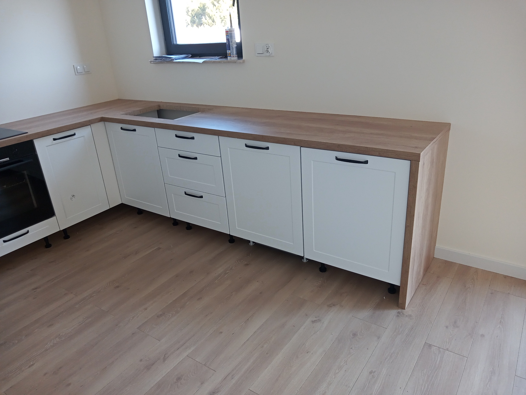 Nowoczesna, jasna kuchnia z białymi szafkami i drewnianym blatem, widoczny zlewozmywak i płyta grzewcza, minimalistyczne uchwyty szafek.