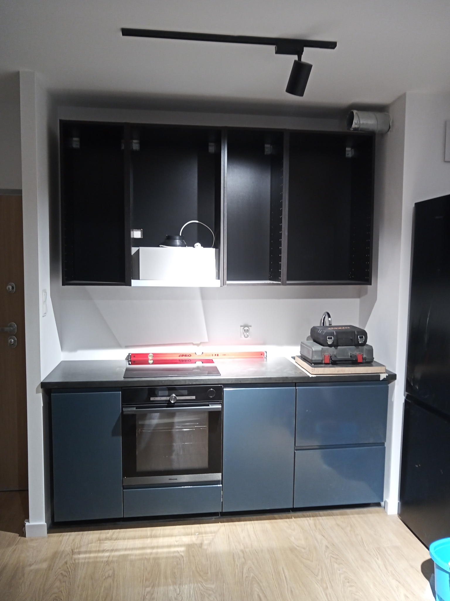 Wnętrze kuchni w trakcie montażu: widoczne wiszące szafki bez frontów, dolne szafki z piekarnikiem w odcieniach szarości i niebieskiego, poziomica na blacie oraz skrzynki narzędziowe. Uwagę zwraca...