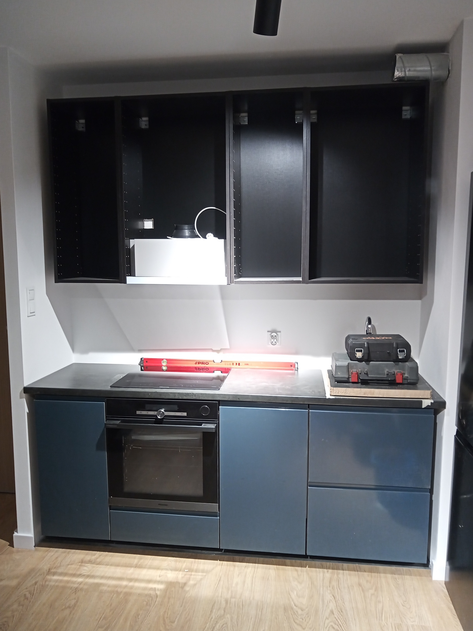 Częściowo zamontowana kuchnia z ciemnymi szafkami wiszącymi bez frontów, niebieskimi szafkami dolnymi, wbudowanym piekarnikiem i poziomnicą na blacie roboczym.