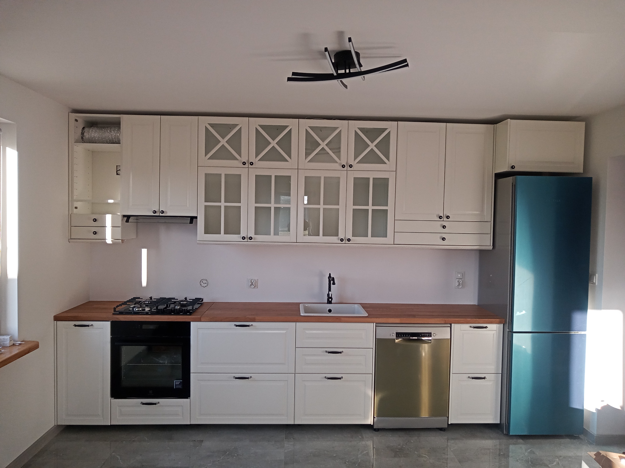 Biała kuchnia z drewnianym blatem, widoczny turkusowy lodówka, piekarnik i płyta gazowa, srebrna zmywarka, oraz nowoczesna lampa sufitowa.