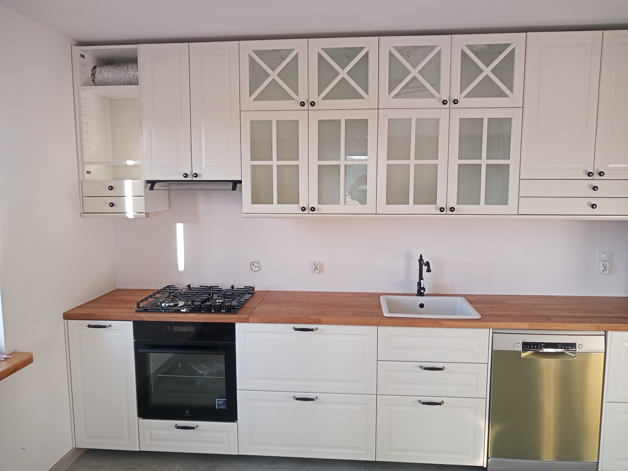 Biała kuchnia z drewnianym blatem, szklanymi frontami szafek wiszących i czarnymi uchwytami, z widoczną kuchenką gazową, piekarnikiem i zmywarką.