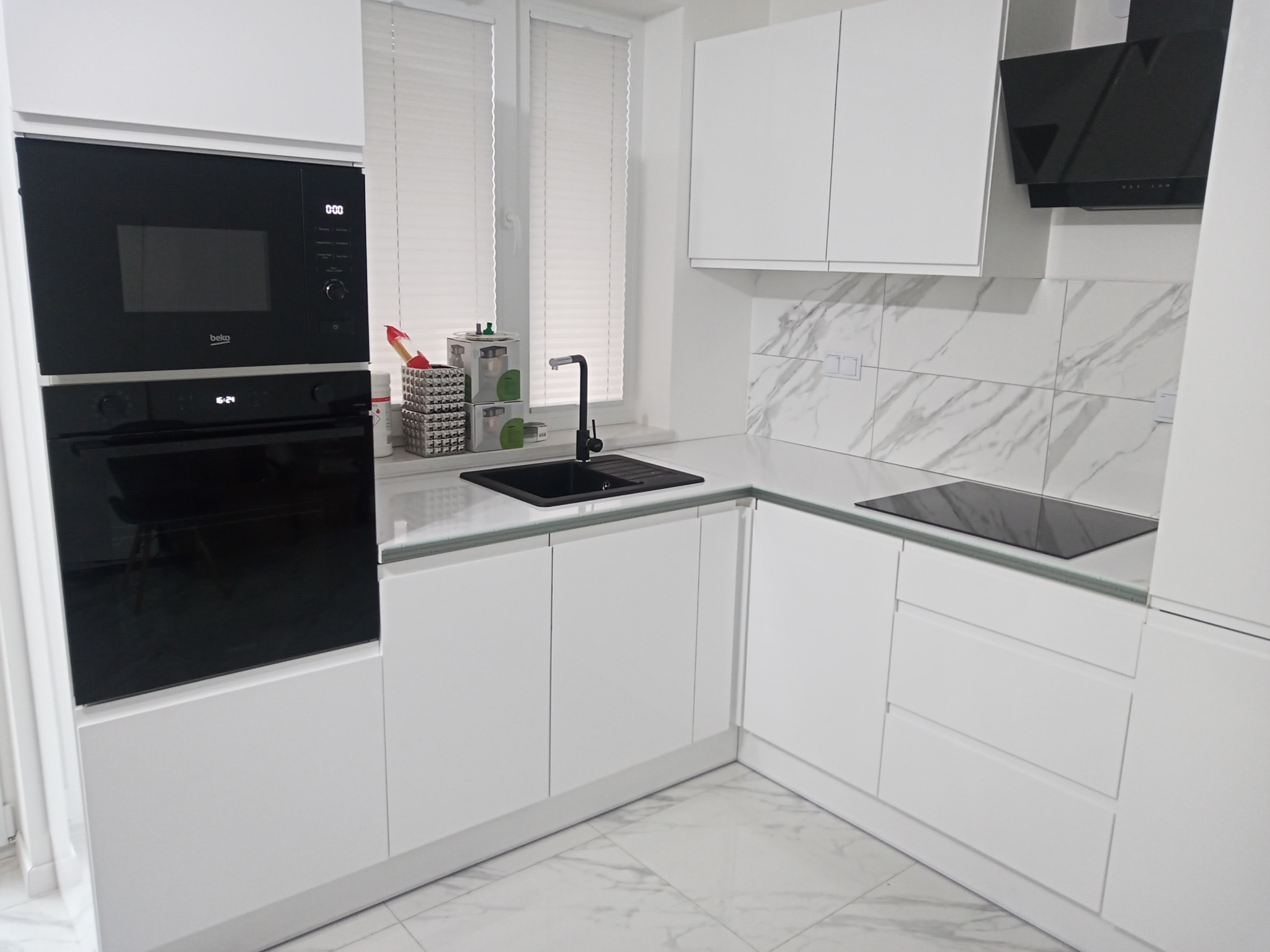 Nowoczesna, minimalistyczna kuchnia z białymi frontami, czarną armaturą i sprzętem AGD, marmurowym wzorem na ścianie nad blatem i podłogą w tym samym stylu.