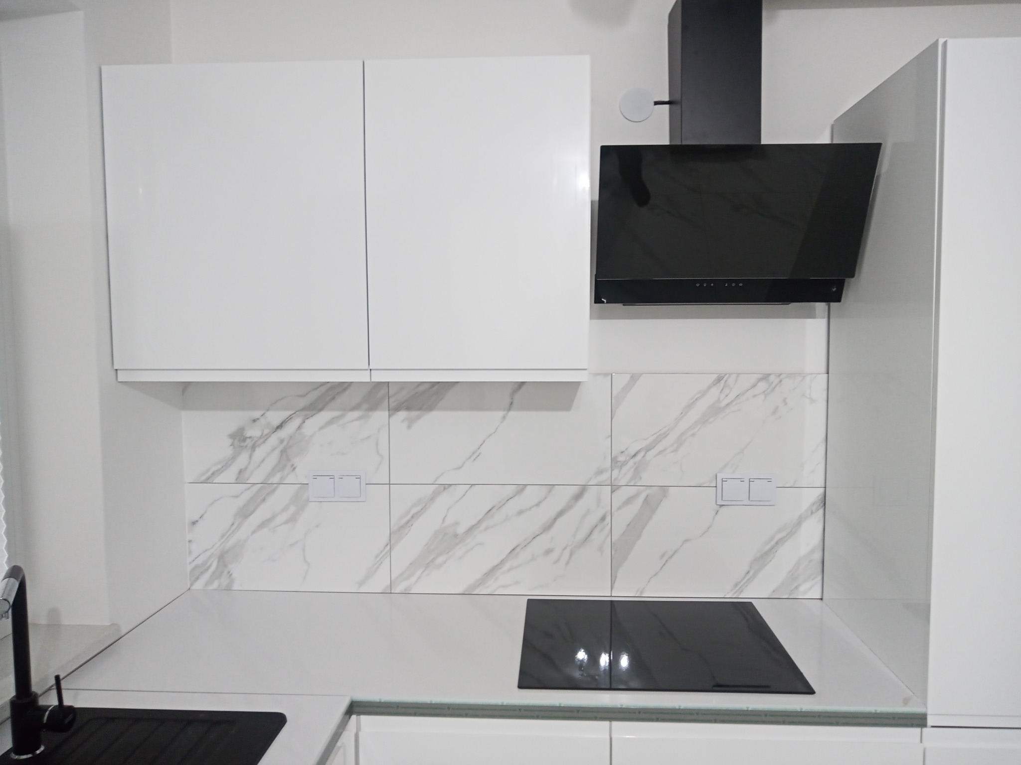 Nowoczesna, minimalistyczna kuchnia z białymi, lakierowanymi szafkami, czarną płytą indukcyjną i okapem, ściana wykończona płytkami imitującymi marmur z szarymi żyłami, widoczna bateria...
