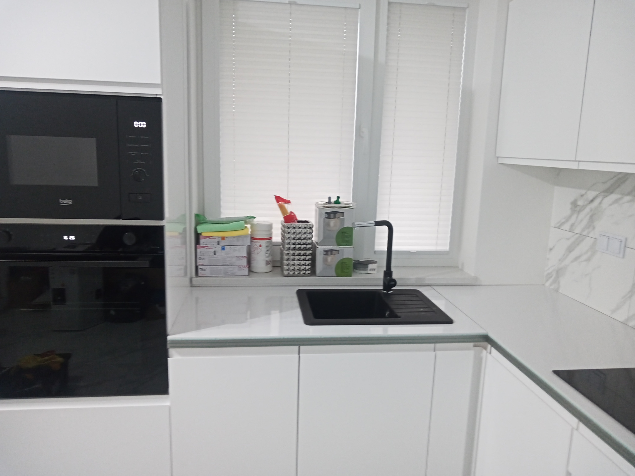 Nowoczesna biała kuchnia z czarnym zlewem i baterią, widok na okno z żaluzjami, zabudowa kuchenna z piekarnikiem i mikrofalówką marki Beko.