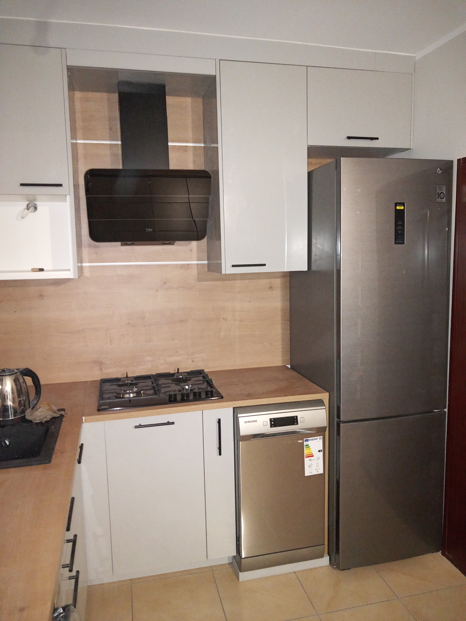 Nowoczesna kuchnia z szarymi szafkami, drewnianym blatem, czarnym okapem i sprzętem AGD: lodówka, zmywarka, płyta gazowa.