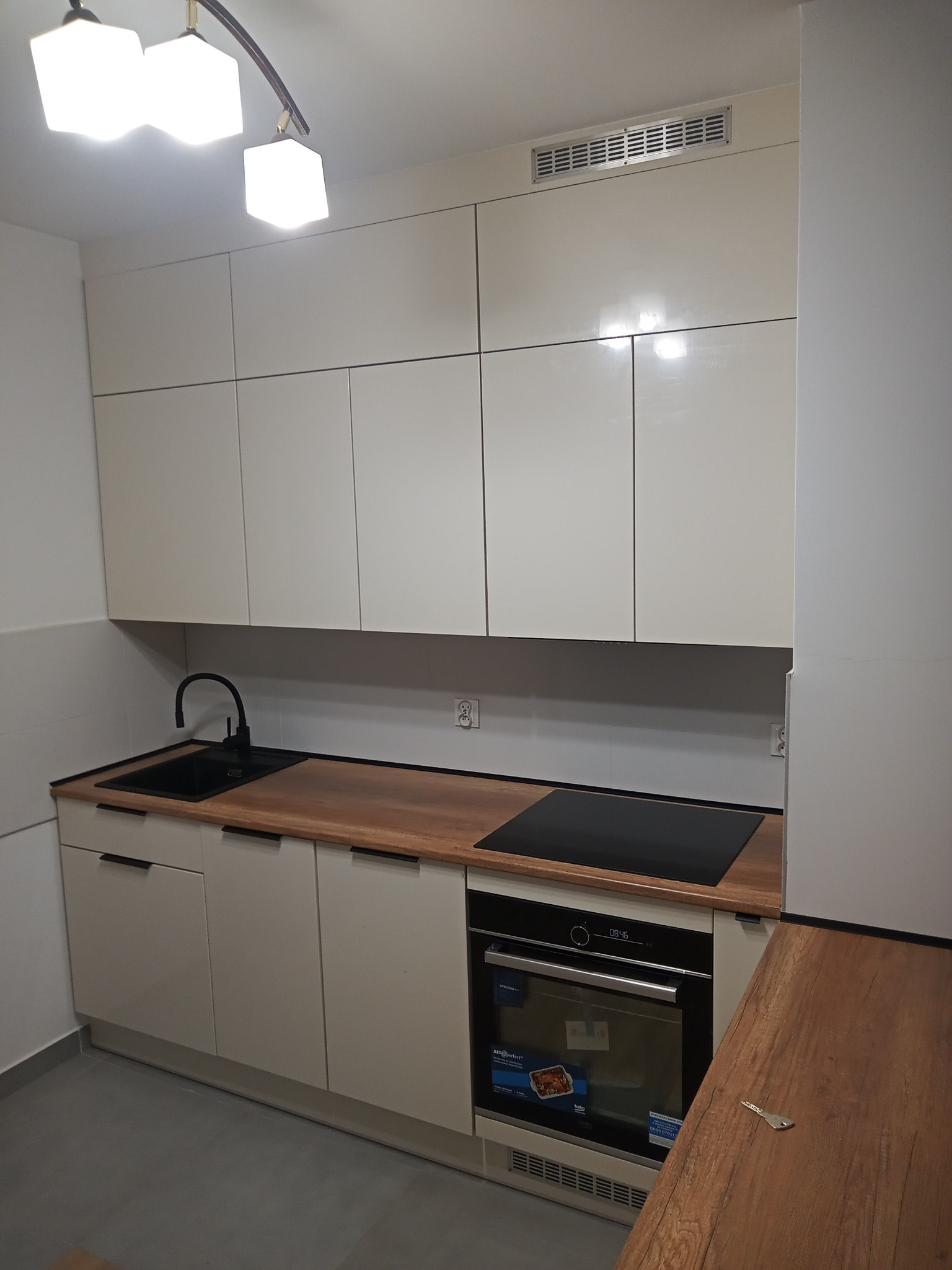 Nowoczesna, jasna kuchnia z górnymi szafkami sięgającymi sufitu, drewnianym blatem, czarnym zlewem i płytą indukcyjną. Na blacie leży pojedynczy klucz.