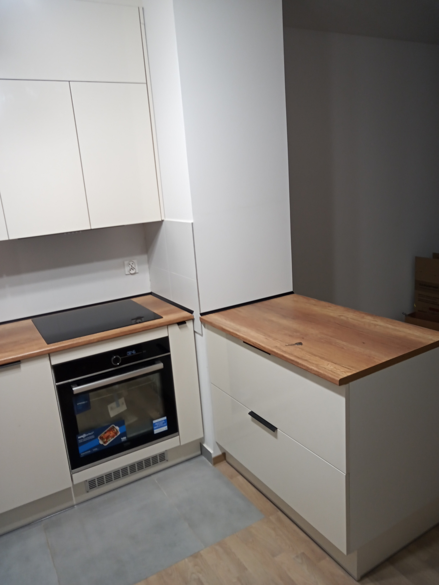 Nowoczesna zabudowa kuchenna z białymi szafkami i drewnianym blatem, widoczny piekarnik z wbudowaną płytą grzewczą.