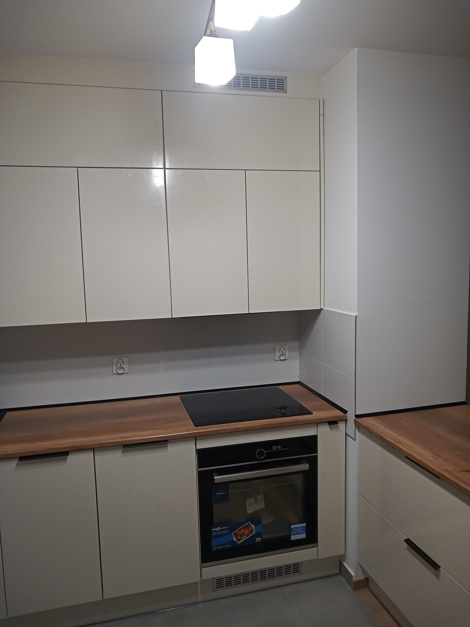 Kremowa zabudowa kuchenna z drewnianym blatem, płyta indukcyjna i piekarnik w zabudowie, widoczne gniazdka elektryczne na ścianie.