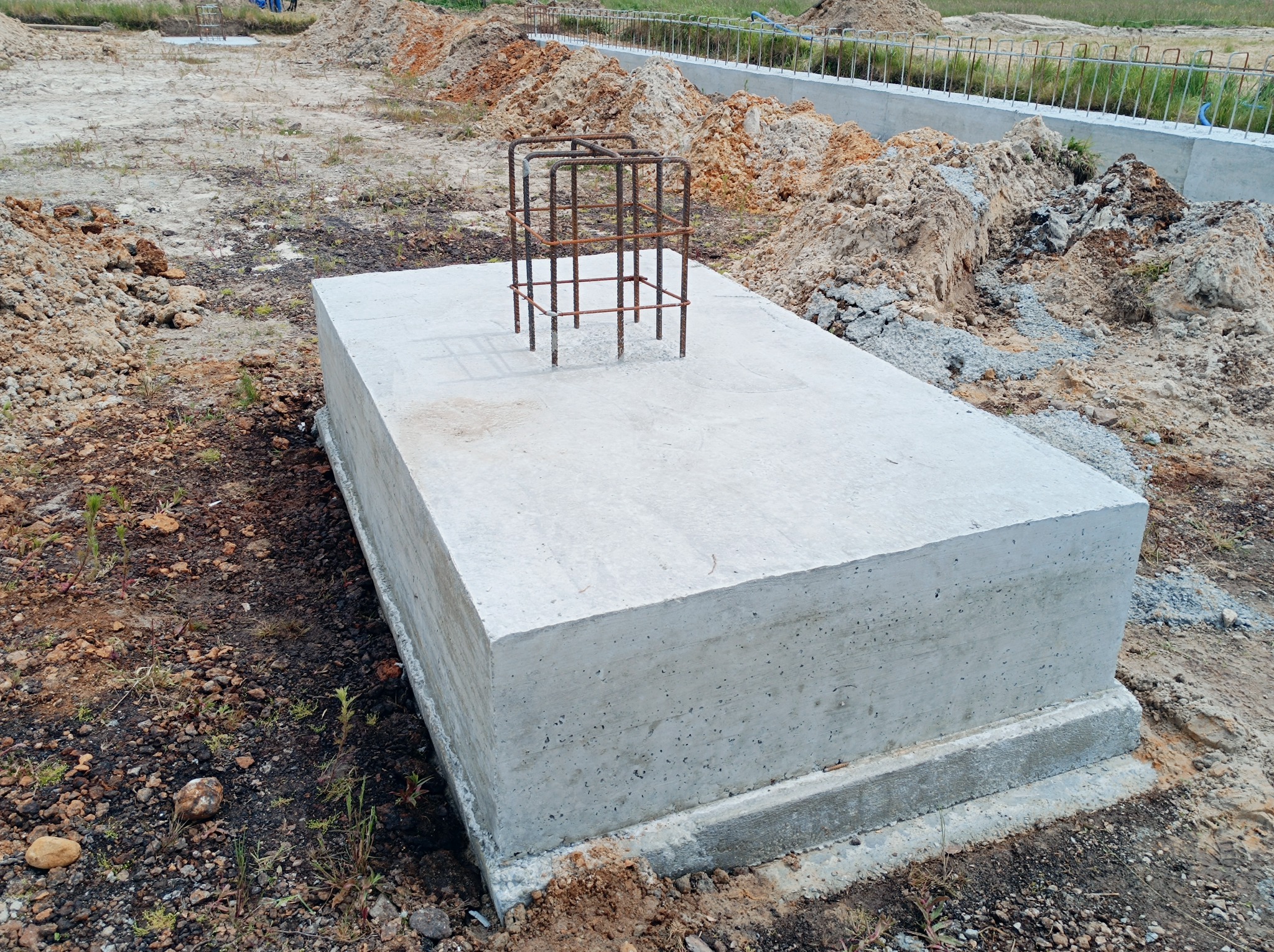Betonowy fundament z wystającą zbrojeniem na placu budowy, otoczony ziemią i gruzem.