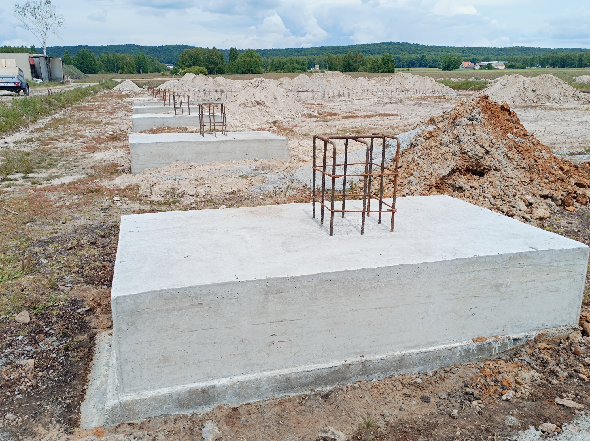 Betonowe bloki fundamentowe z wystającymi prętami zbrojeniowymi, widok na plac budowy z hałdami ziemi i odległym lasem.