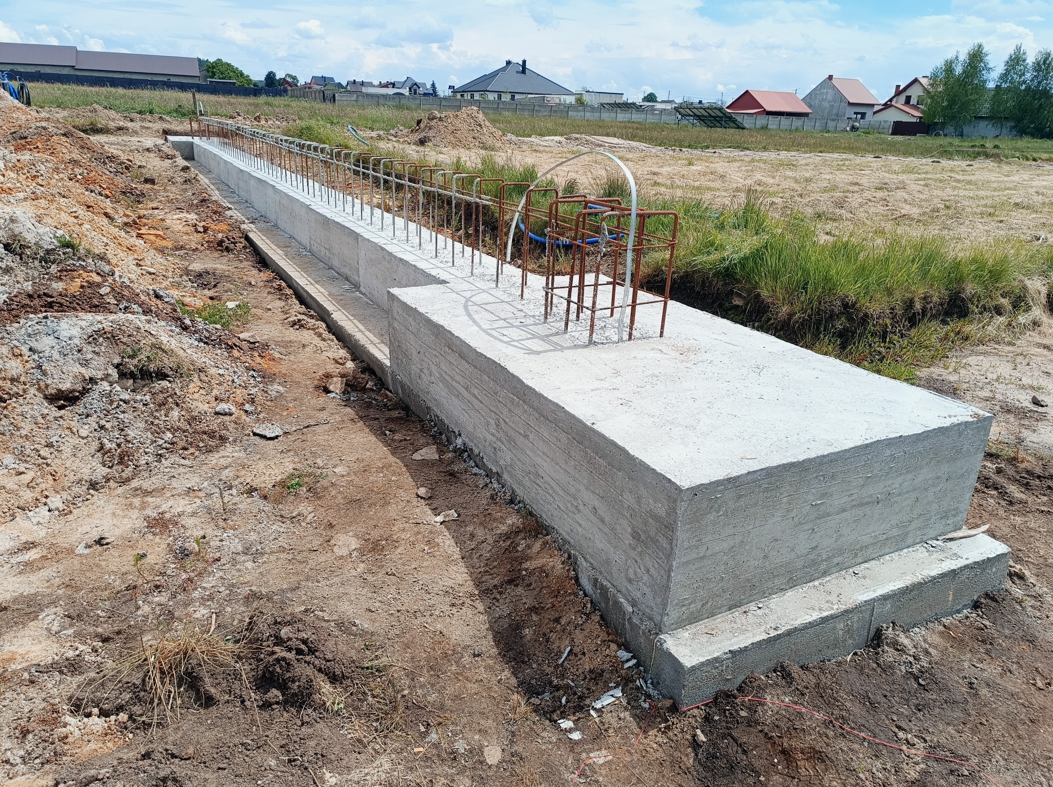 Betonowy fundament z wystającymi prętami zbrojeniowymi na budowie domu, widok z boku, w tle domy jednorodzinne.