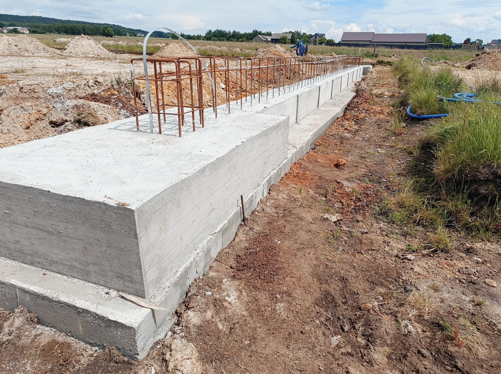 Wylane betonowe ławy fundamentowe z wystającą stalową konstrukcją zbrojeniową, ziemia wokół, w tle budynki mieszkalne.