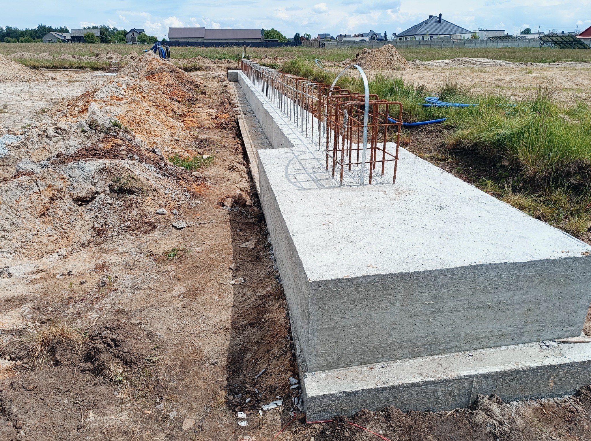 Betonowy fundament z wystającymi prętami zbrojeniowymi na placu budowy, widoczne ślady po szalunku, w tle domy jednorodzinne.