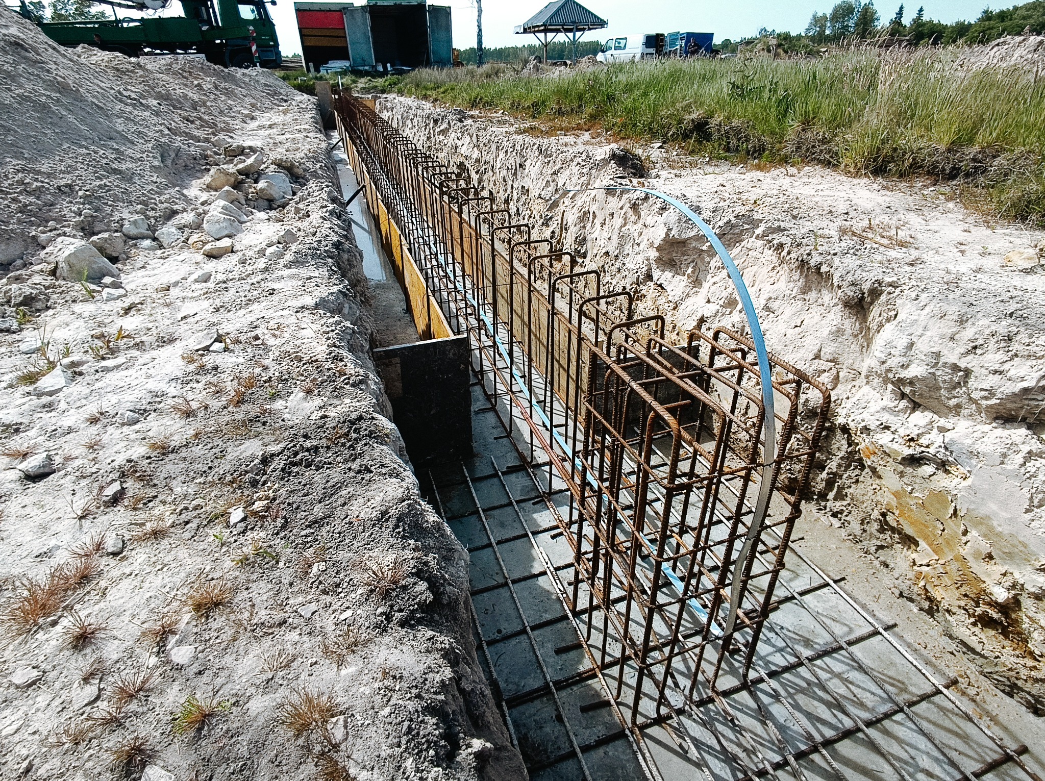 Zbrojenie szalunku pod wylewkę fundamentu w wykopie, widoczna metalowa siatka i pionowe pręty zbrojeniowe połączone niebieską rurką, piasek i ziemia po bokach wykopu, w tle zieleń i budynki...