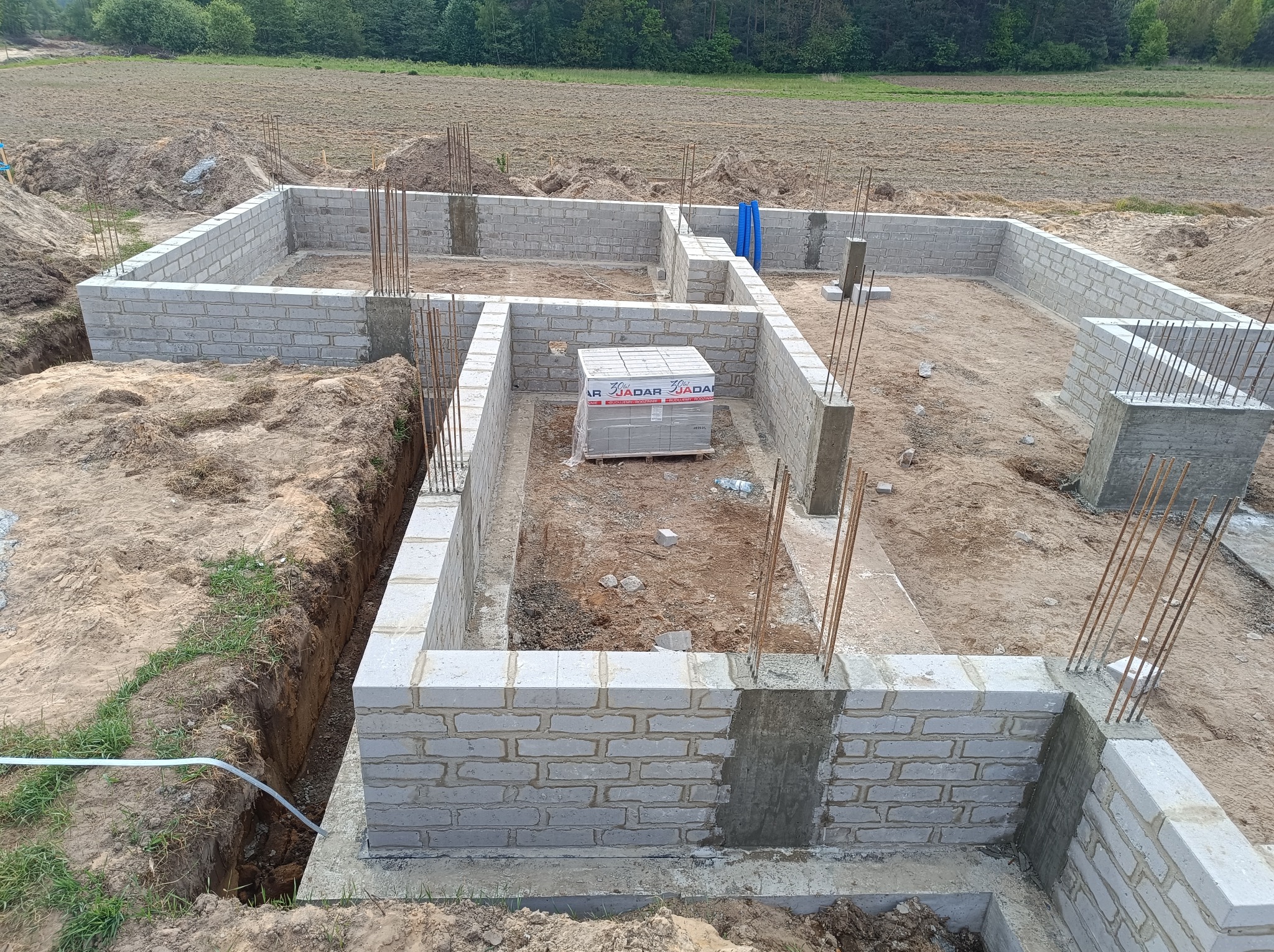 Wykopane fundamenty z częściowo wymurowanymi ścianami z bloczków betonowych, wystające pręty zbrojeniowe, paleta bloczków wewnątrz, widok z góry na plac budowy.
