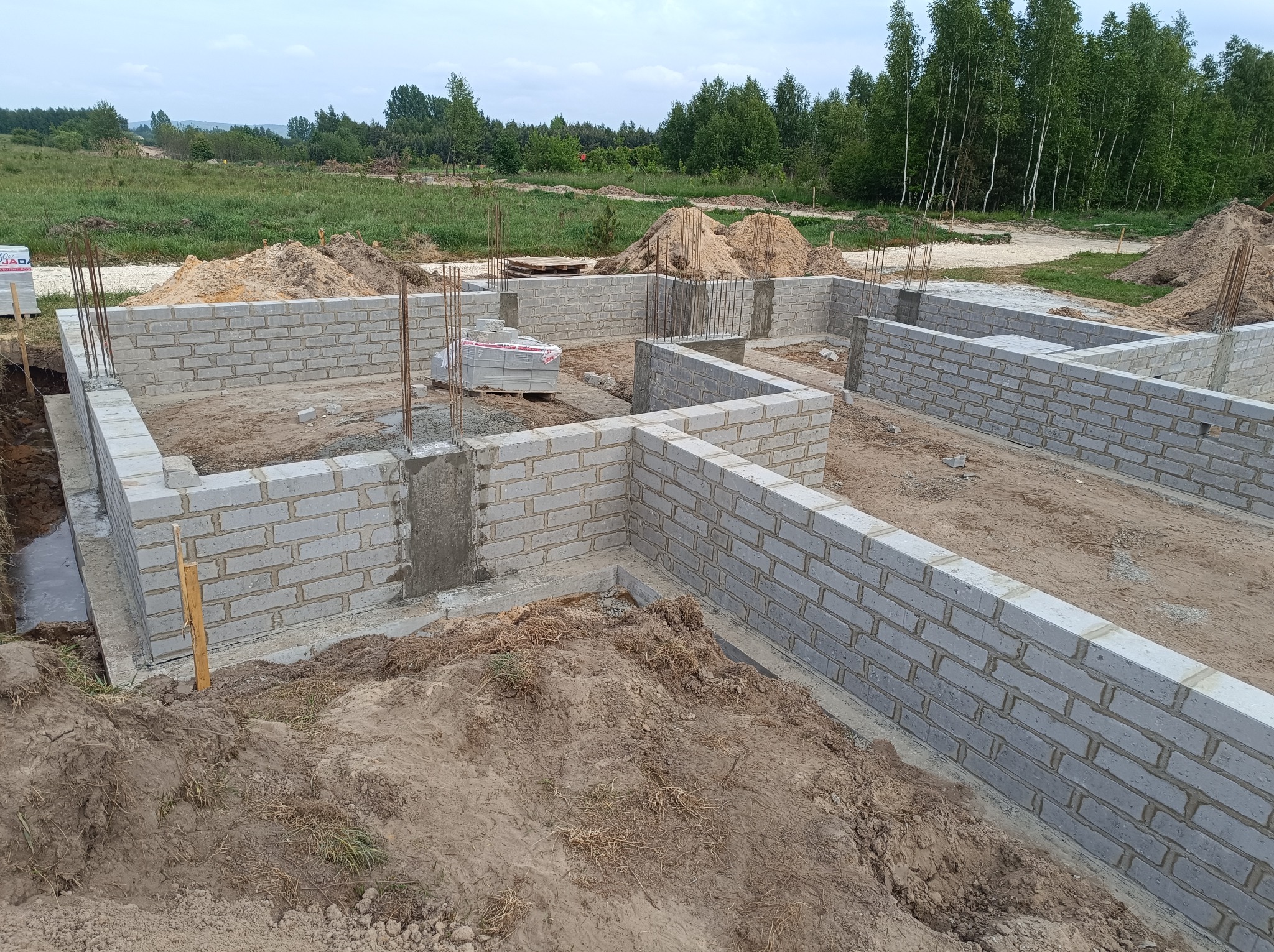 Wznoszone mury fundamentowe z bloczków betonowych na tle krajobrazu z drzewami i hałdami ziemi.