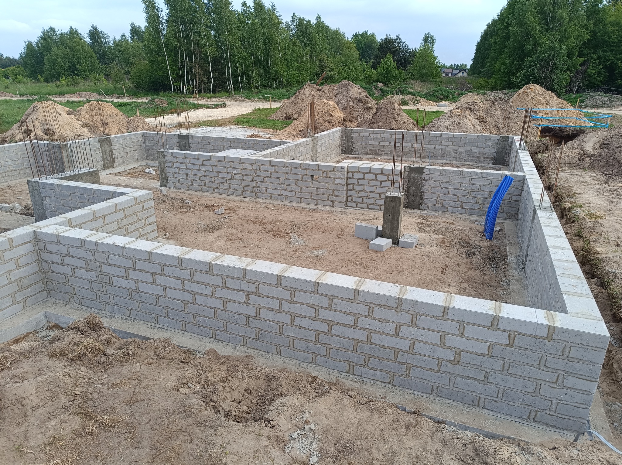 Wzniesione ściany fundamentowe z bloczków betonowych, widoczne pręty zbrojeniowe, hałdy piasku w tle, niebieska rura na krawędzi wykopu.