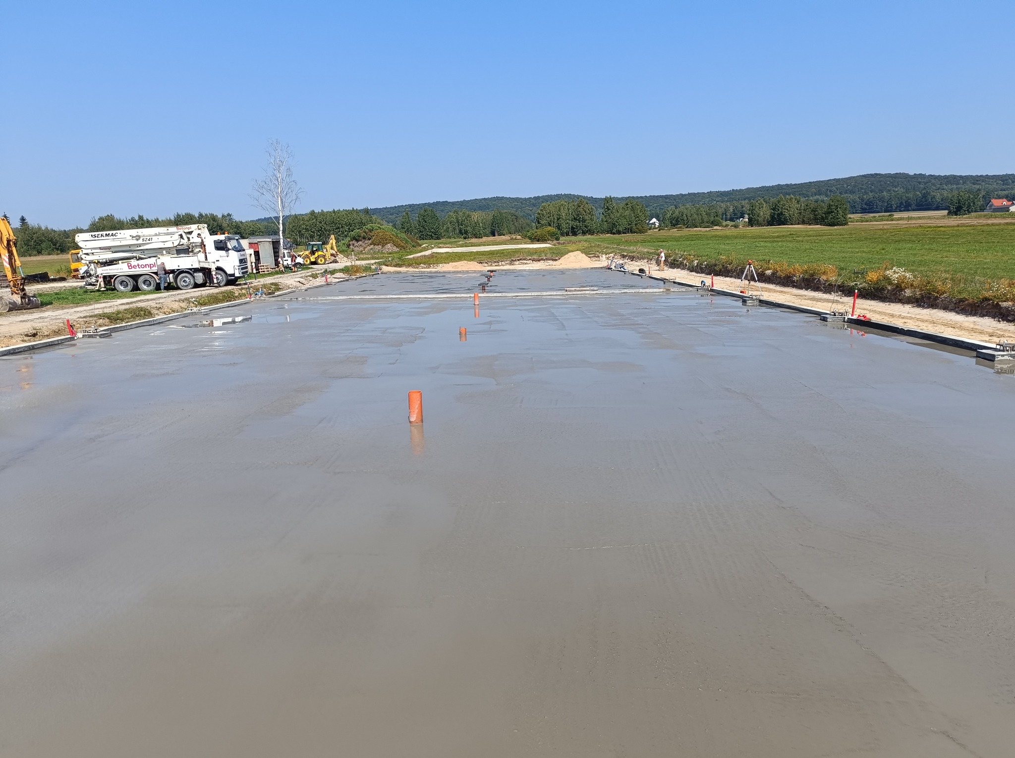 Świeżo wylana, gładka płyta fundamentowa z widocznymi pomarańczowymi rurkami pionowymi, z betonomieszarką i sprzętem budowlanym w tle, na tle zielonego krajobrazu.