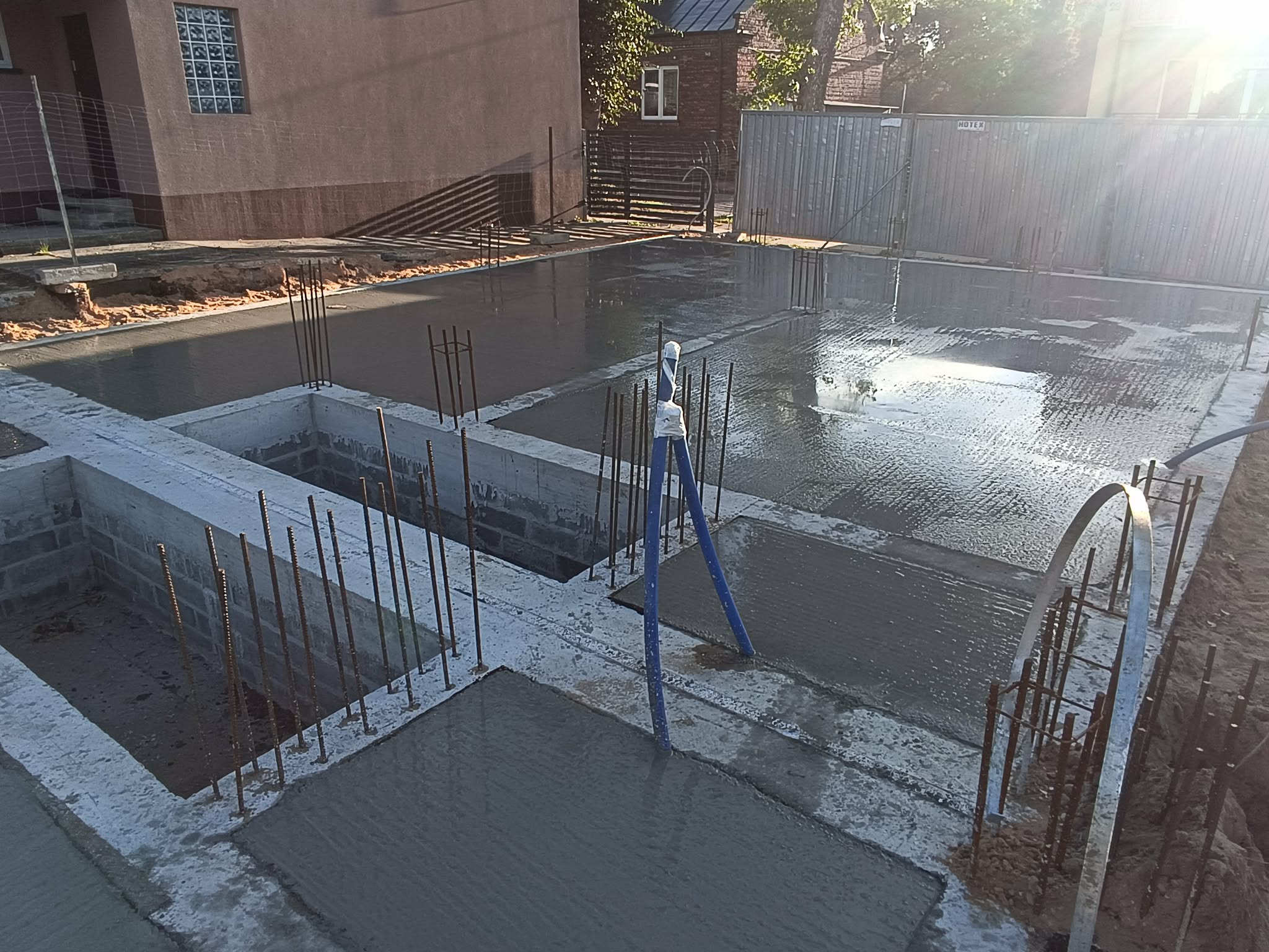 Zalane betonem fundamenty z wystającymi prętami zbrojeniowymi, widoczne bloczki fundamentowe i szalunki. W tle budynek i ogrodzenie.