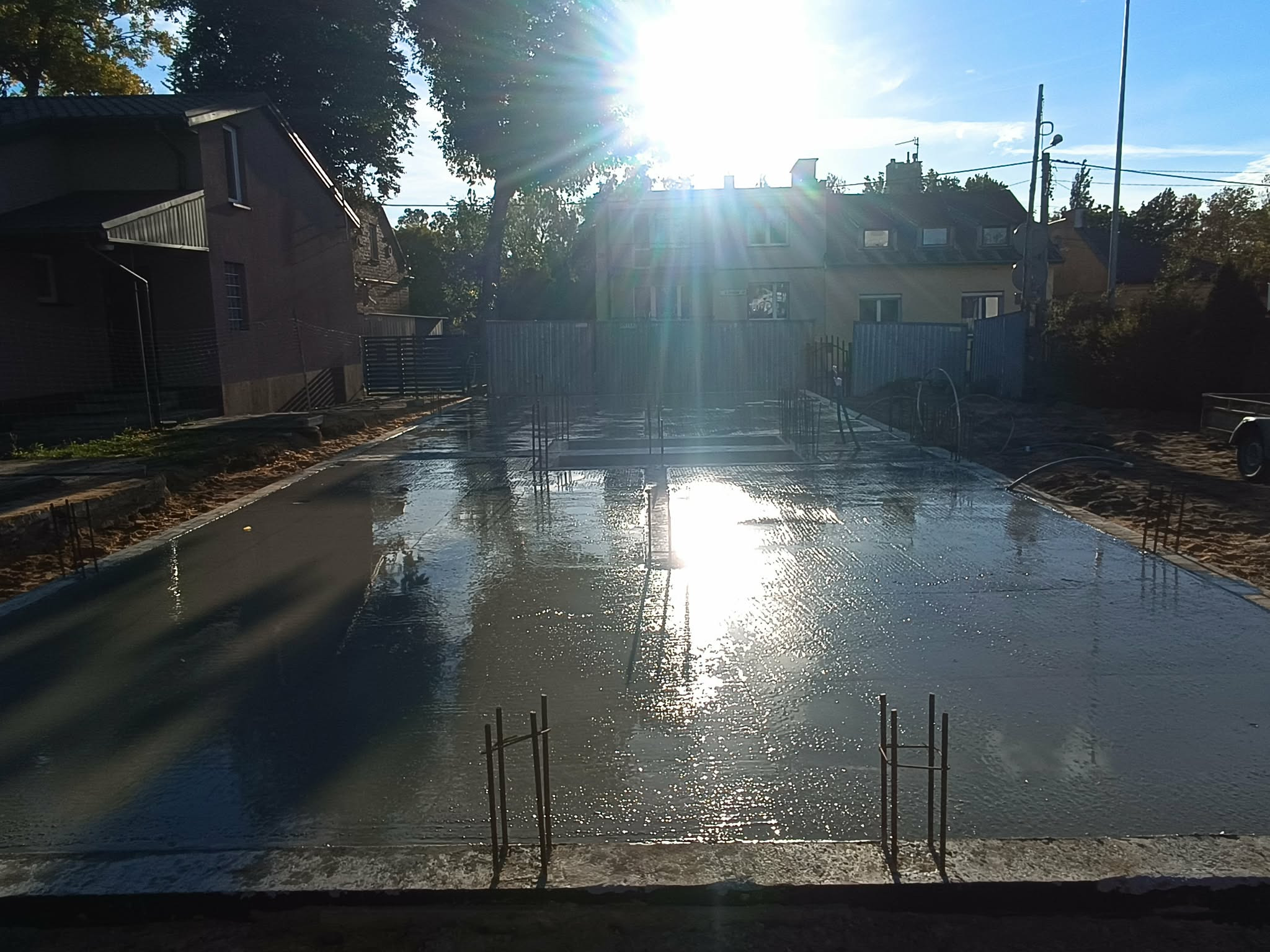 Zalany fundament pod budynek w słoneczny dzień. Widoczne wystające pręty zbrojeniowe i odbicia słońca w mokrym betonie. W tle budynki mieszkalne i ogrodzenie.