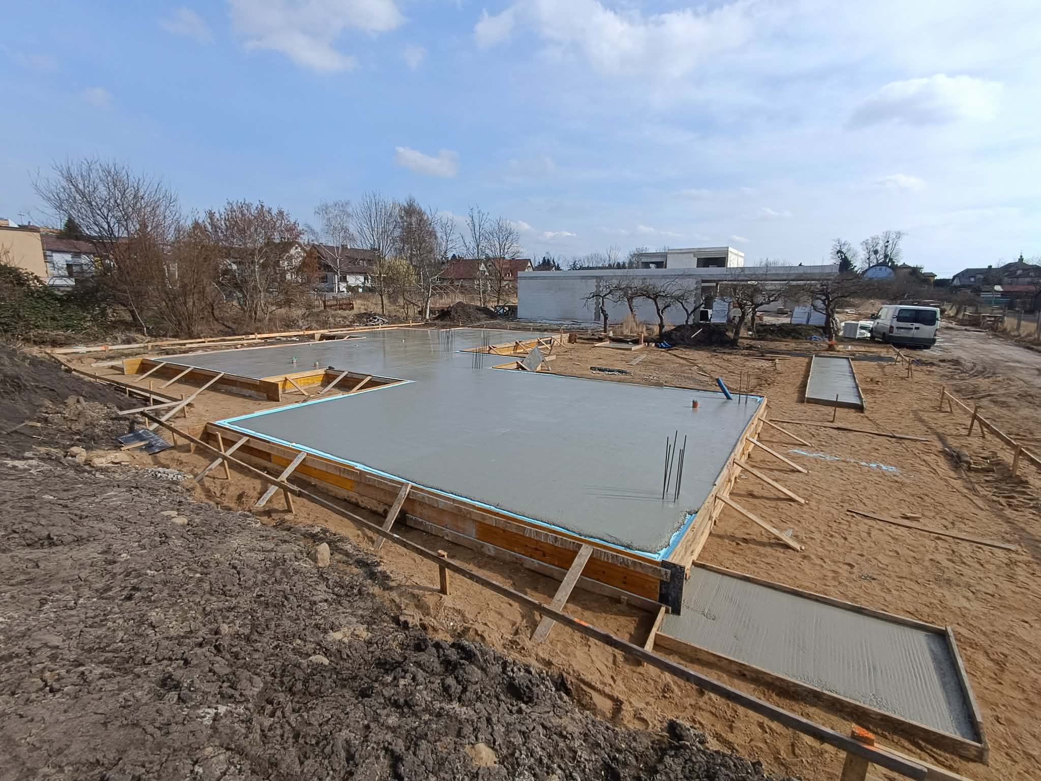 Wylany fundament z drewnianym szalunkiem i izolacją, widok z góry na plac budowy w słoneczny dzień. W tle zabudowa jednorodzinna i błękitne niebo.