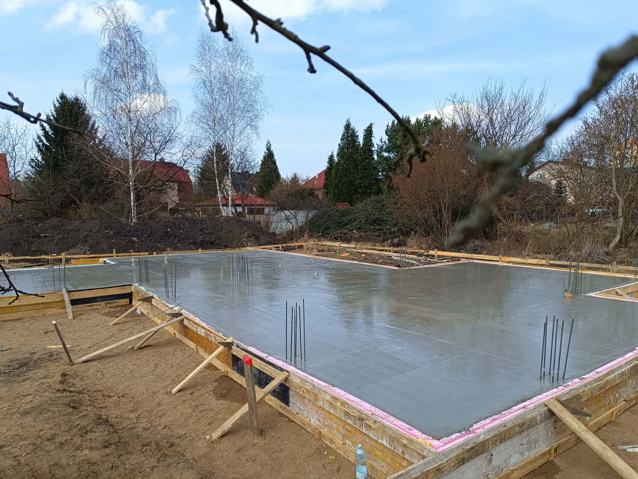 Zalany fundament pod dom jednorodzinny, z widocznym szalunkiem i wystającymi prętami zbrojeniowymi. W tle drzewa i zabudowania. Ujęcie z poziomu gruntu.