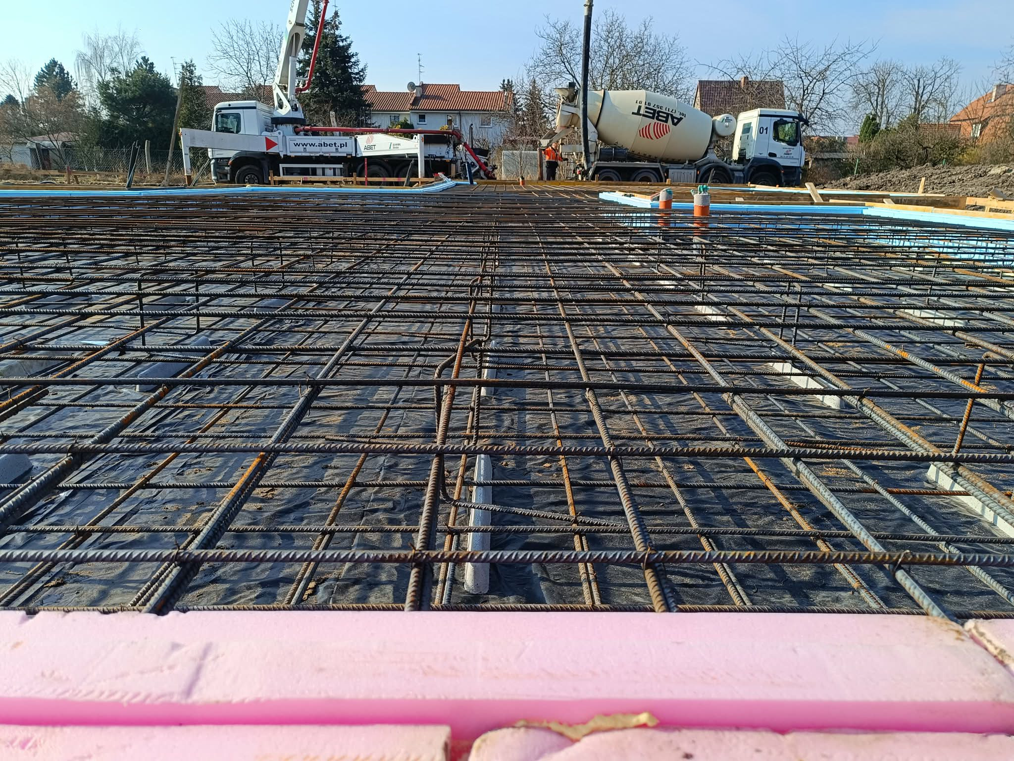 Zbrojenie fundamentów z widocznymi prętami, folią i izolacją termiczną. W tle betoniarki i budynki w trakcie budowy. Perspektywa z dołu, podkreślająca strukturę zbrojenia.