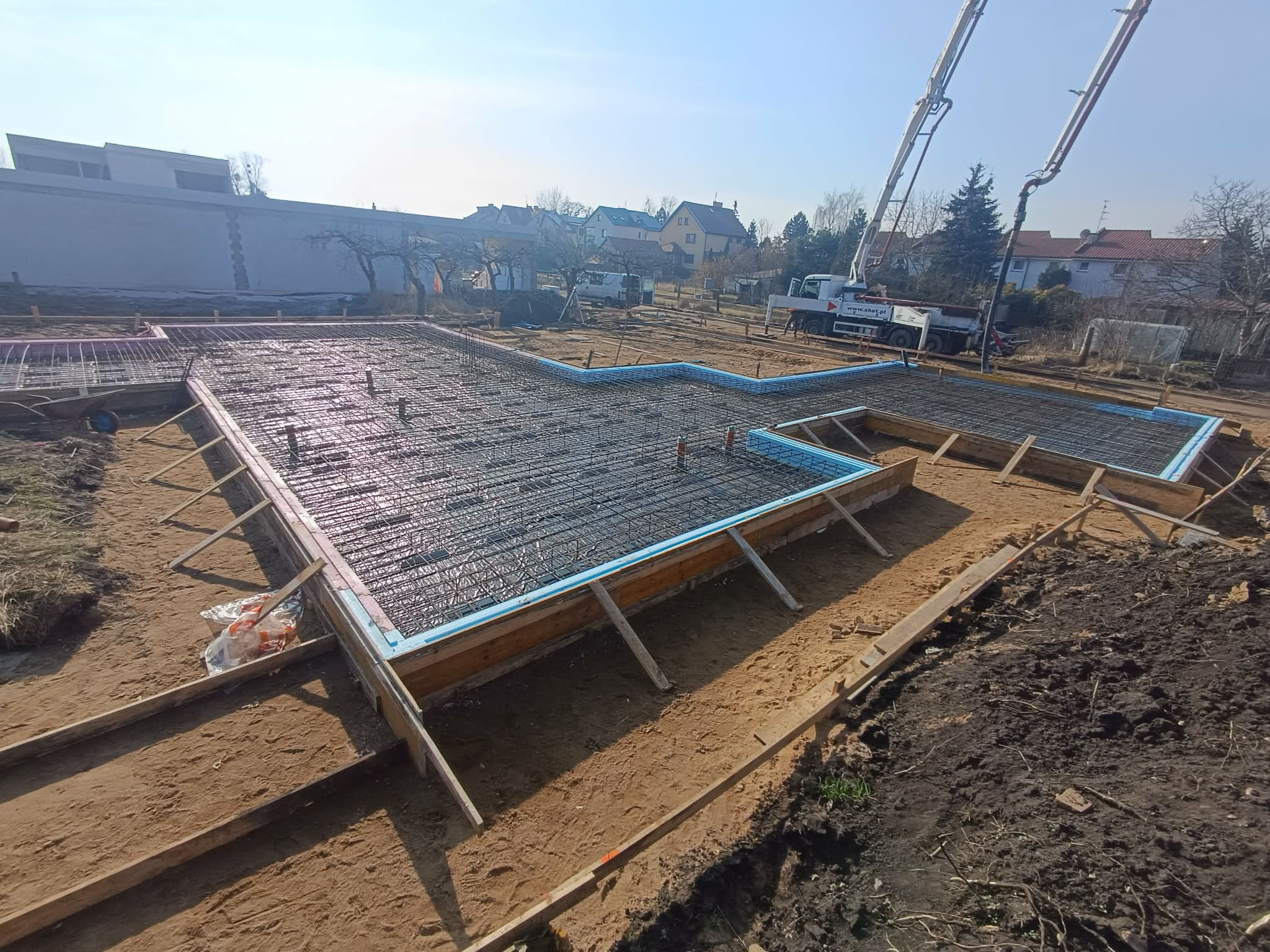 Zbrojenie fundamentów domu jednorodzinnego z użyciem siatki stalowej i szalunków z izolacją, na tle pompy do betonu i zabudowy jednorodzinnej w słoneczny dzień.