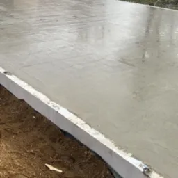 Świeżo wylany, gładki fundament betonowy z białym obrzeżem, otoczony ziemią, w tle trawa i drzewa.