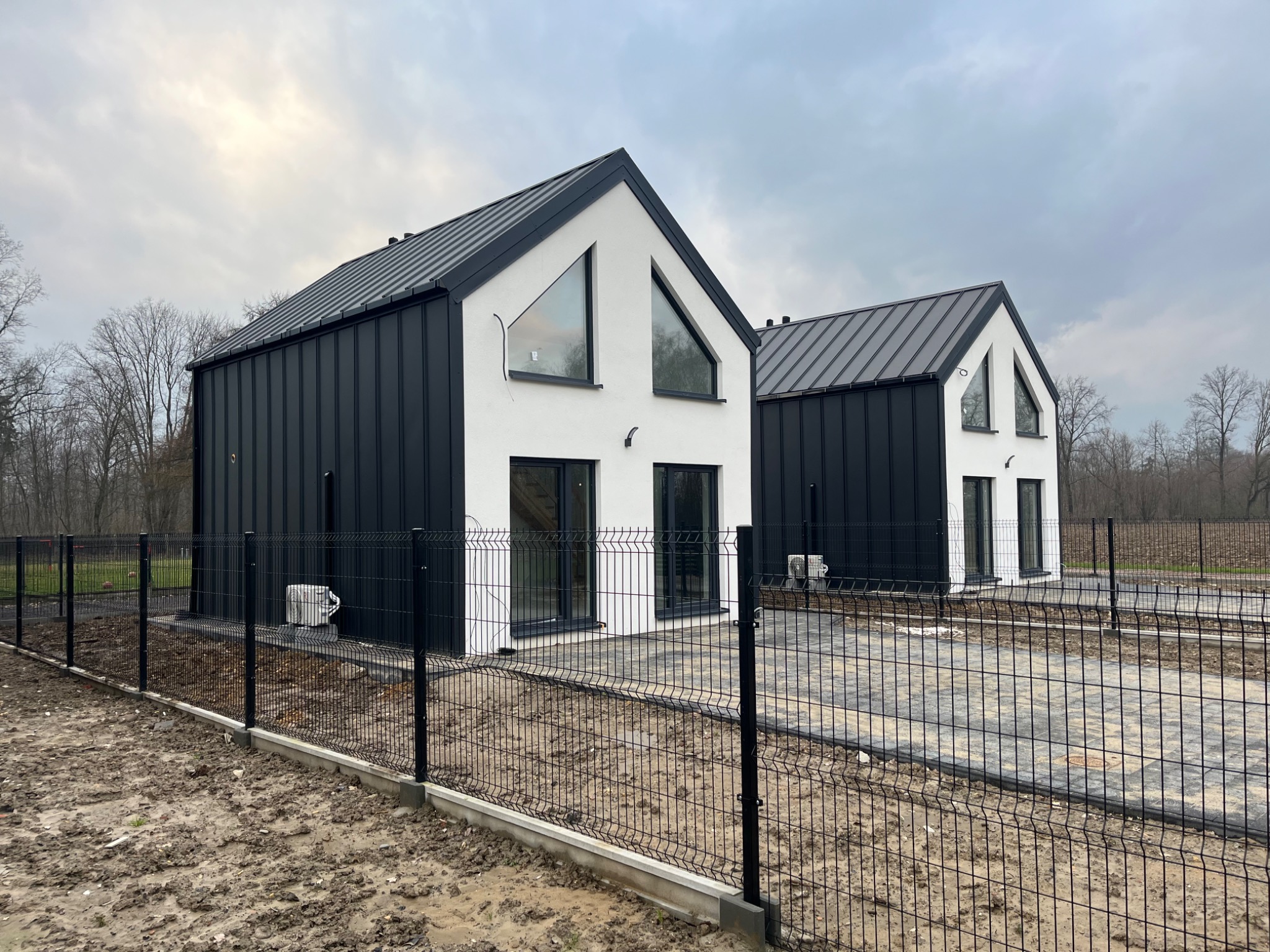 Dwa nowoczesne domy jednorodzinne z czarno-białą elewacją i skośnymi dachami, widoczne jednostki zewnętrzne klimatyzacji na ścianach, otoczone czarnym metalowym ogrodzeniem panelowym na tle...