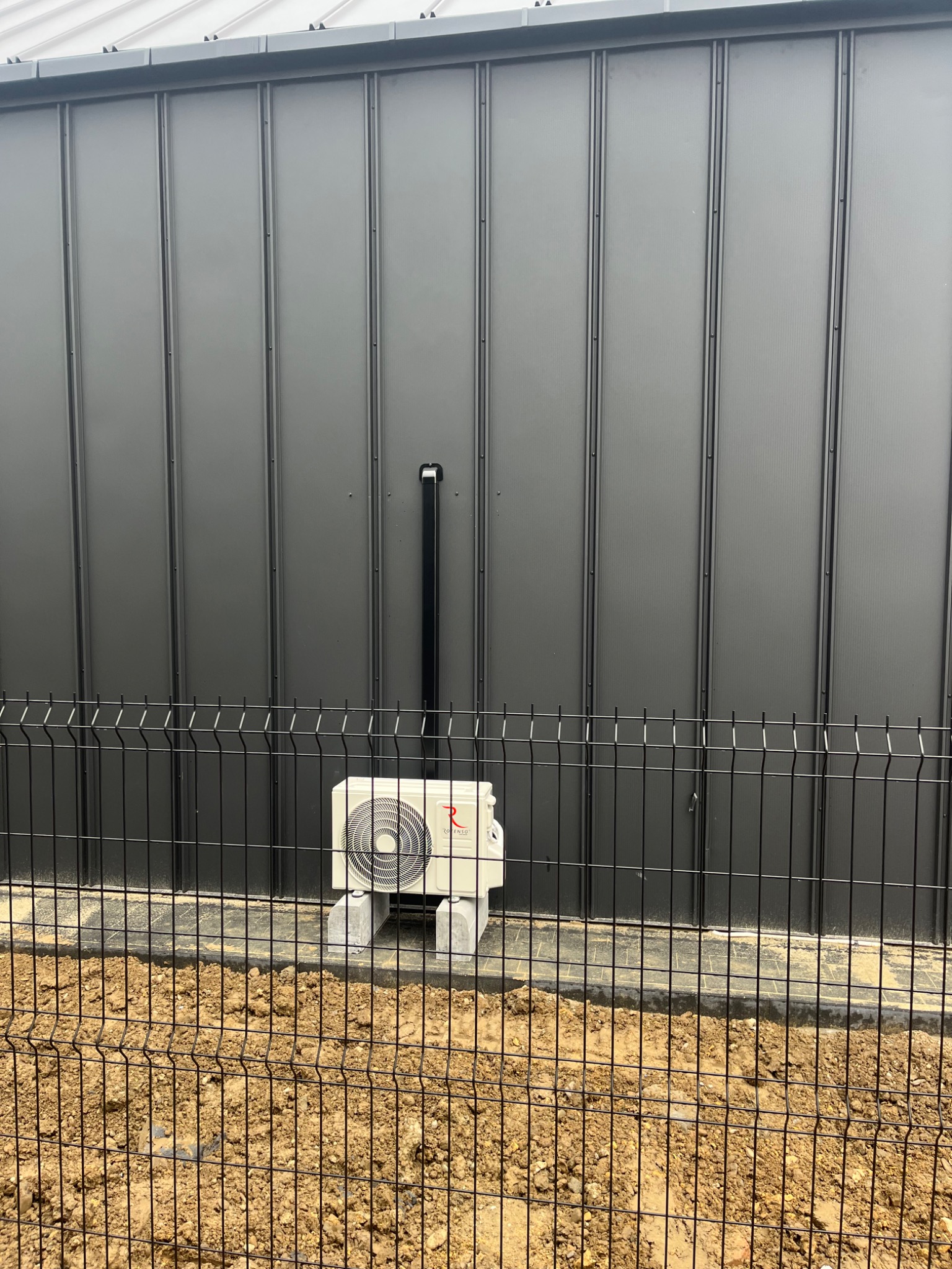 Zewnętrzna jednostka klimatyzacji marki Rotenso umieszczona na betonowych bloczkach przed ciemnoszarą ścianą z paneli, widoczna przez metalowe ogrodzenie.