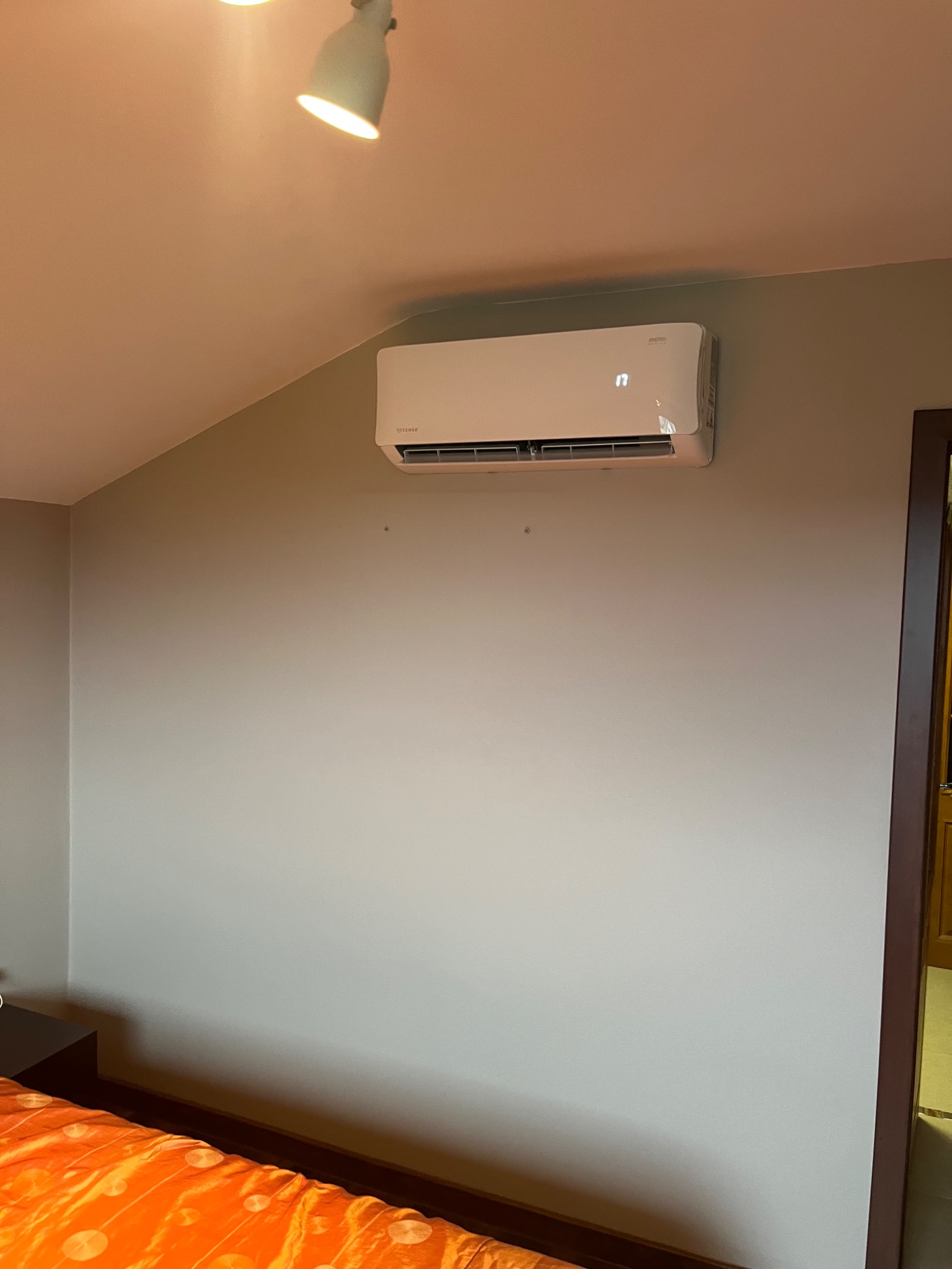 Biały klimatyzator ścienny zamontowany w pokoju na poddaszu, widoczny fragment pomarańczowej pościeli i lampa sufitowa.
