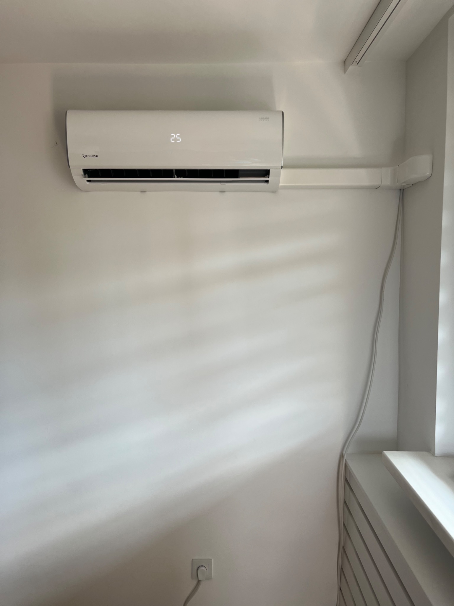 Biała klimatyzacja Rotenso zamontowana na jasnej ścianie, widoczna instalacja elektryczna w listwie, temperatura na wyświetlaczu 25 stopni Celsjusza.