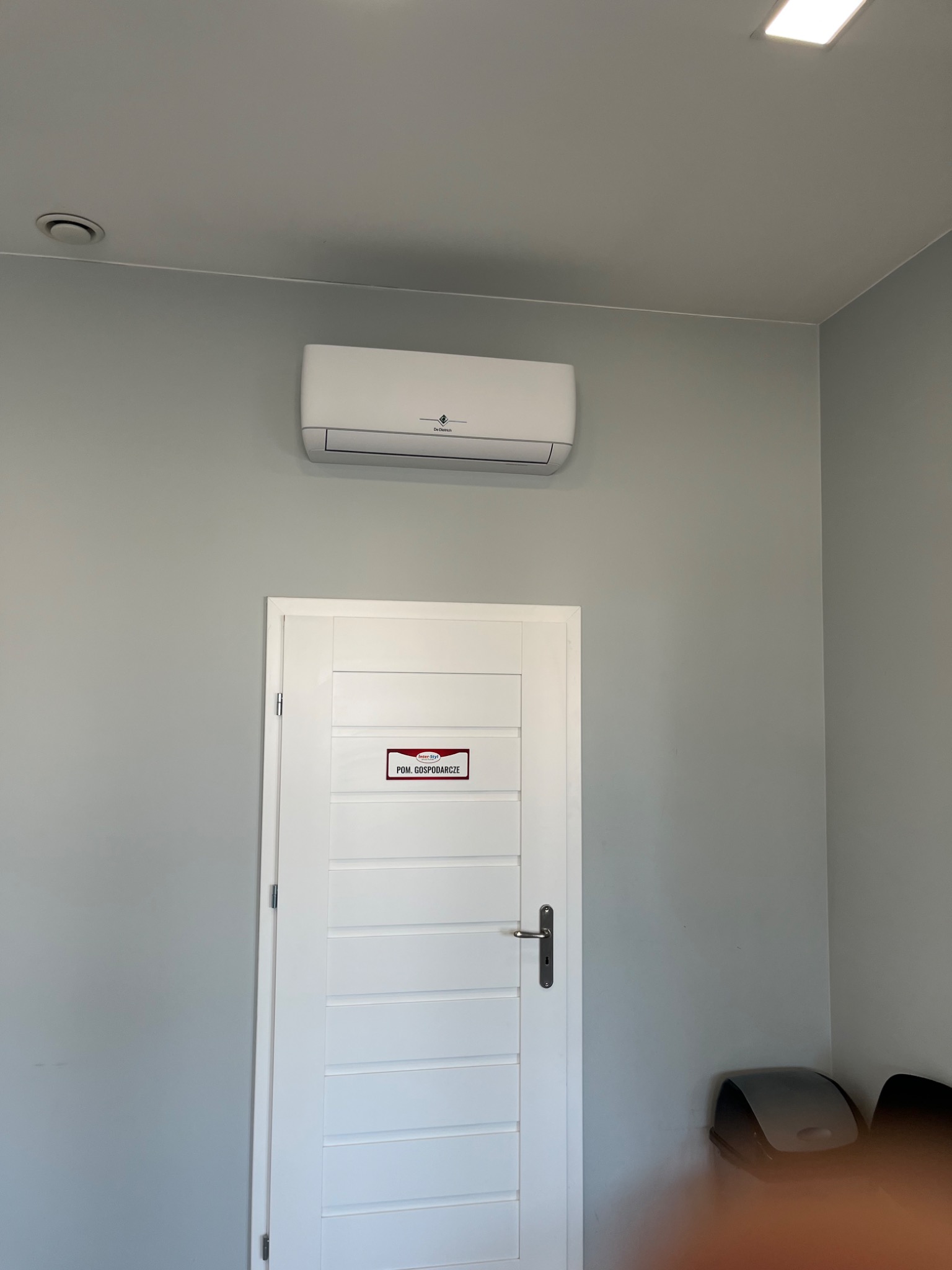 Biały klimatyzator marki De Dietrich zamontowany nad białą, panelową, zamkniętą  drzwi z tabliczką 'Pom. Gospodarcze' w szarym pomieszczeniu.