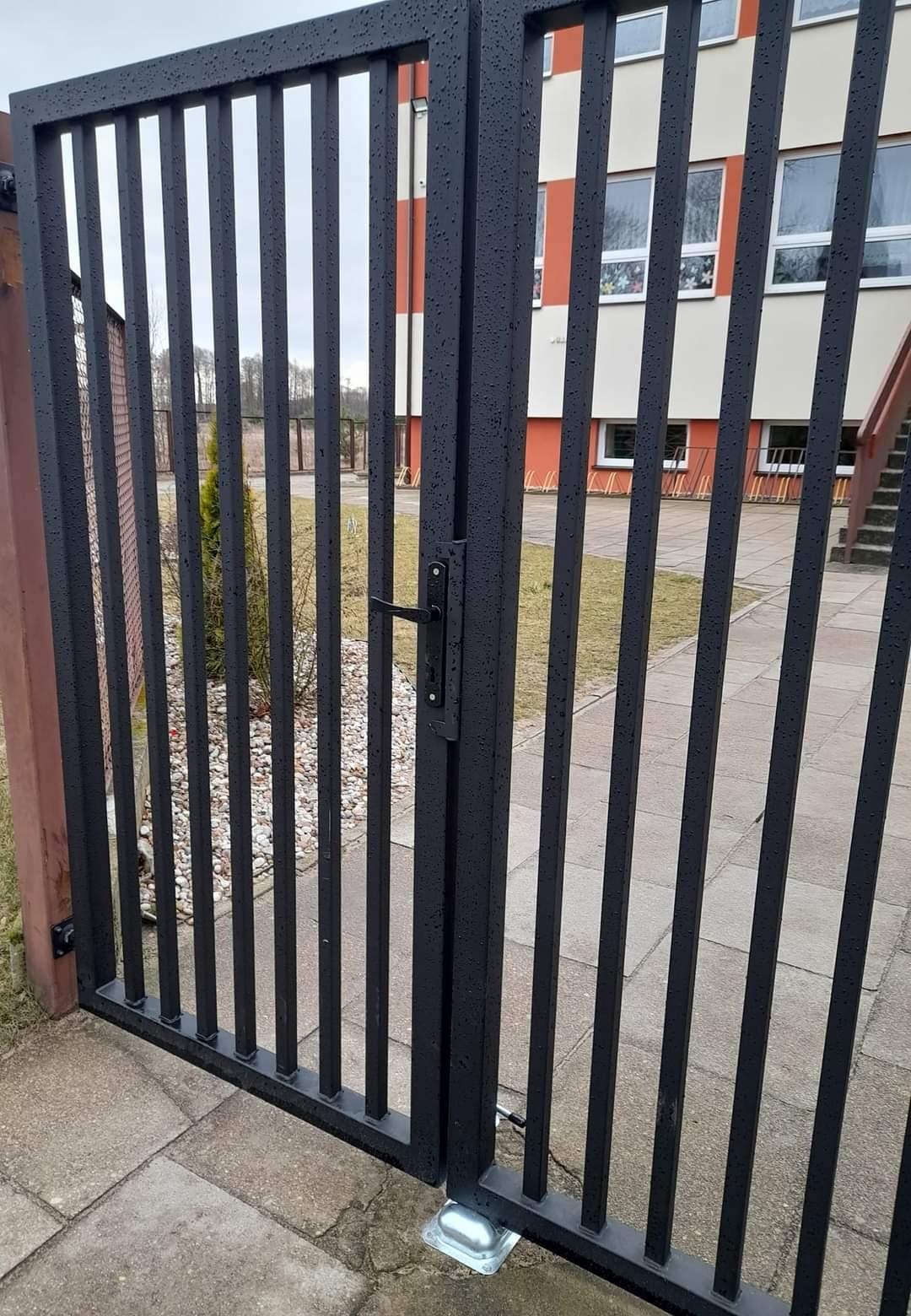 Czarna, metalowa brama wjazdowa z pionowymi szczebelkami, zamontowana na betonowej kostce brukowej, widoczny budynek w tle.