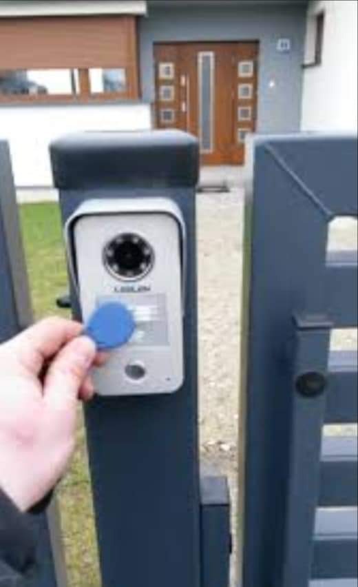 Użytkownik zbliża niebieski transponder do czytnika domofonu zamontowanego na słupku ogrodzenia w kolorze grafitowym, brama uchylna w tle.