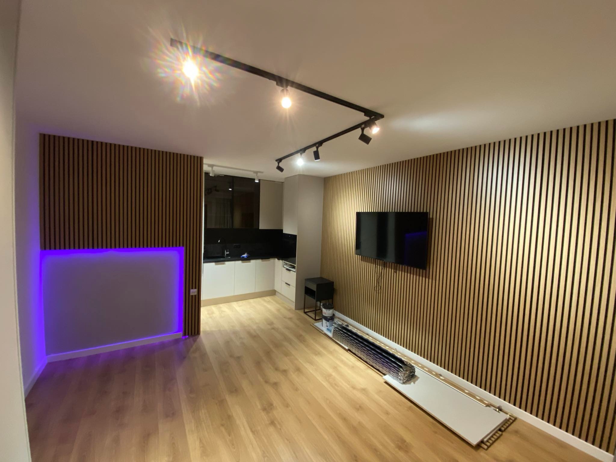 Nowoczesna kawalerka z drewnianymi panelami ściennymi, oświetleniem szynowym i podświetlanym na fioletowo barem, z widoczną otwartą kuchnią w tle, w trakcie prac wykończeniowych.