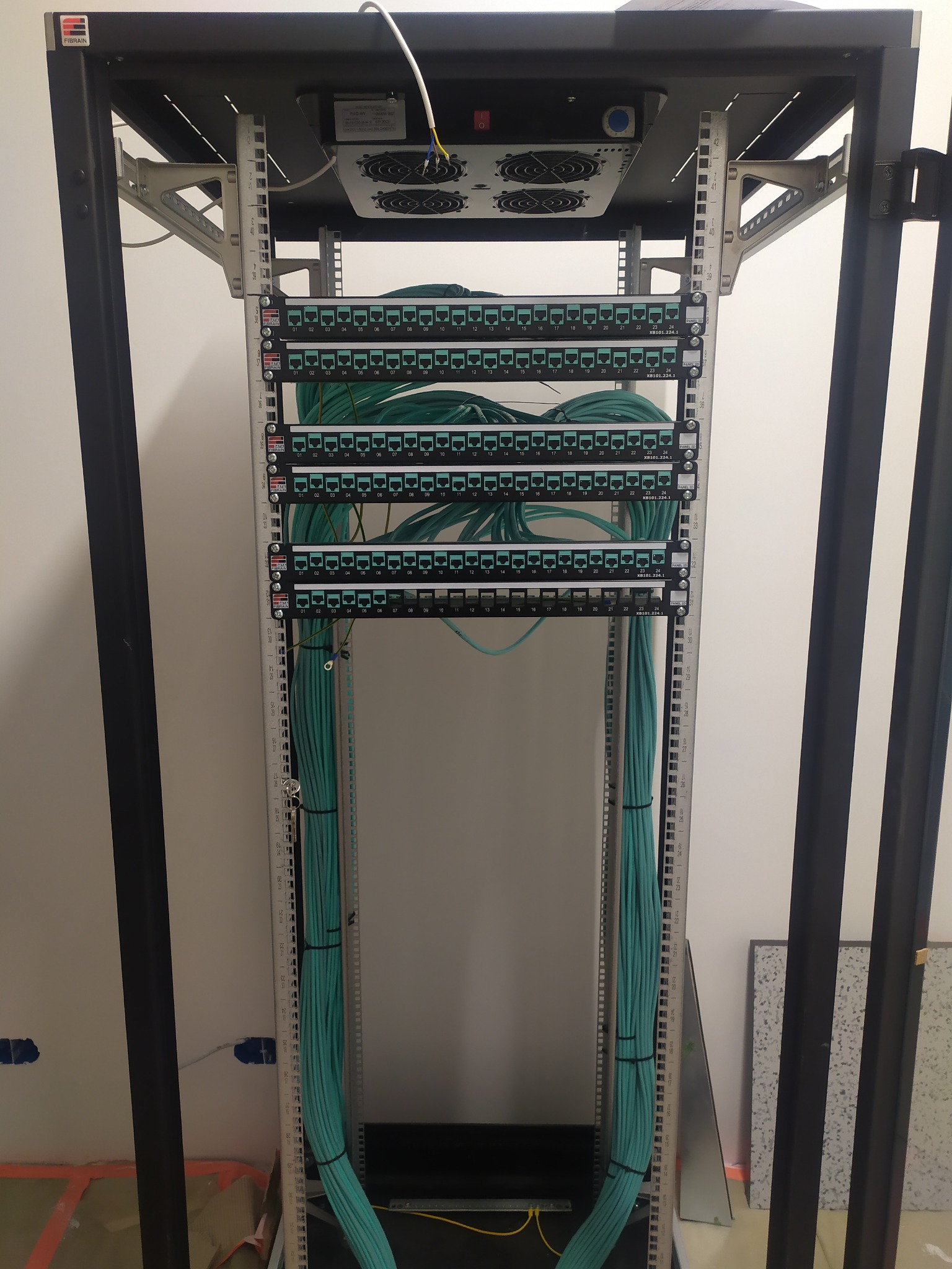 Szafa rack z zamontowanymi panelami krosowymi i uporządkowanymi kablami sieciowymi w kolorze turkusowym, widoczne numery na panelach i oznaczenia portów, wentylatory chłodzące na górze szafy.