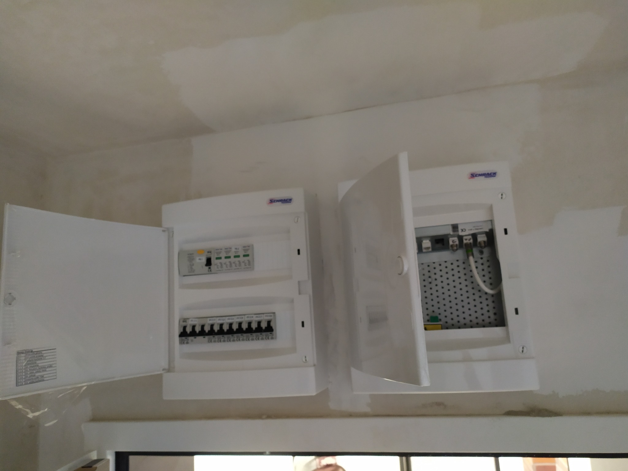 Dwa białe panele elektryczne firmy Schrack zamontowane na ścianie, jeden z otwartymi drzwiczkami ukazującymi wnętrze z okablowaniem i perforowaną płytą montażową, drugi z widocznymi bezpiecznikami...