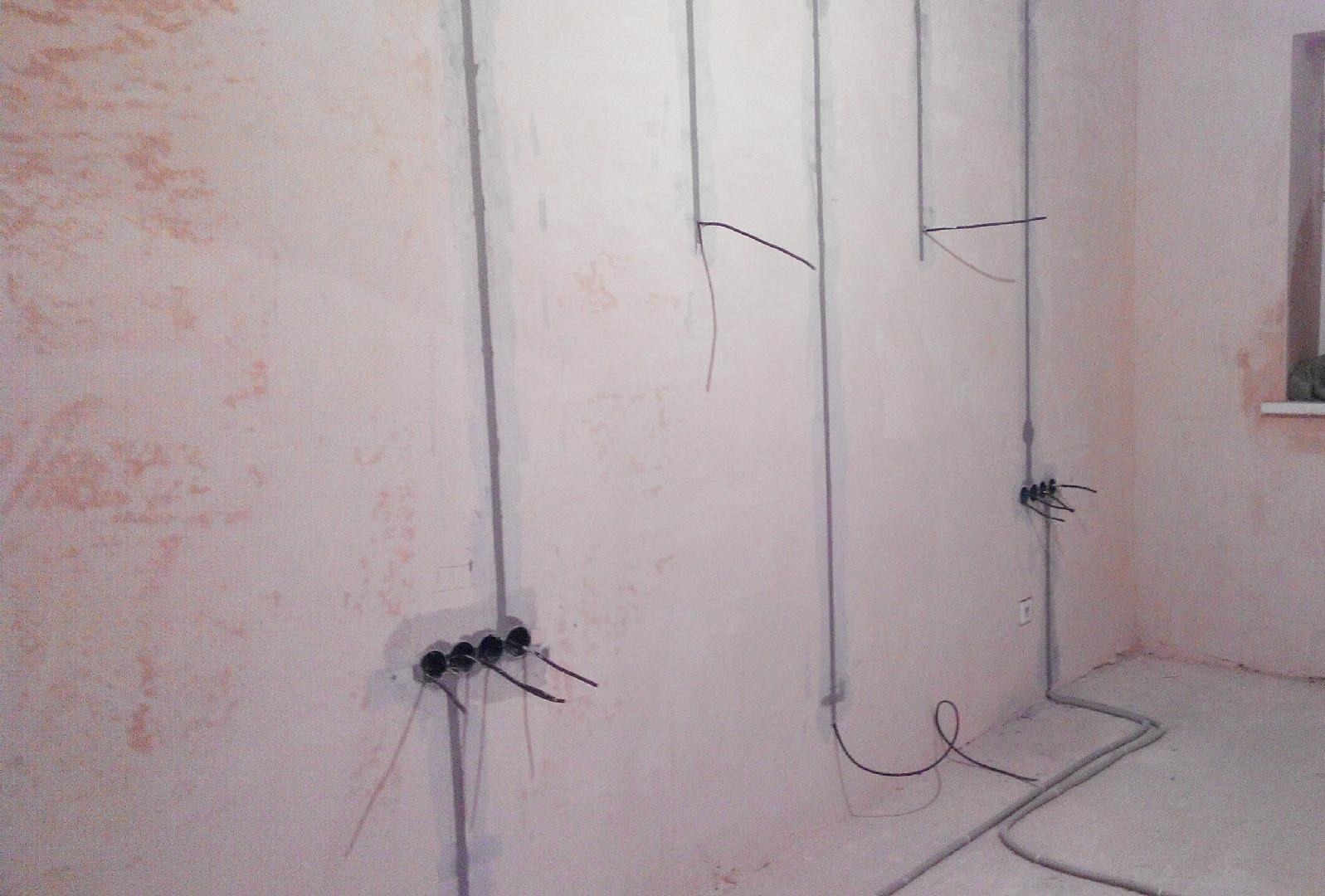 Ściana w trakcie prac elektrycznych: pionowe korytka kablowe, puszki instalacyjne z wystającymi przewodami, oraz peszel na podłodze w surowym pomieszczeniu.