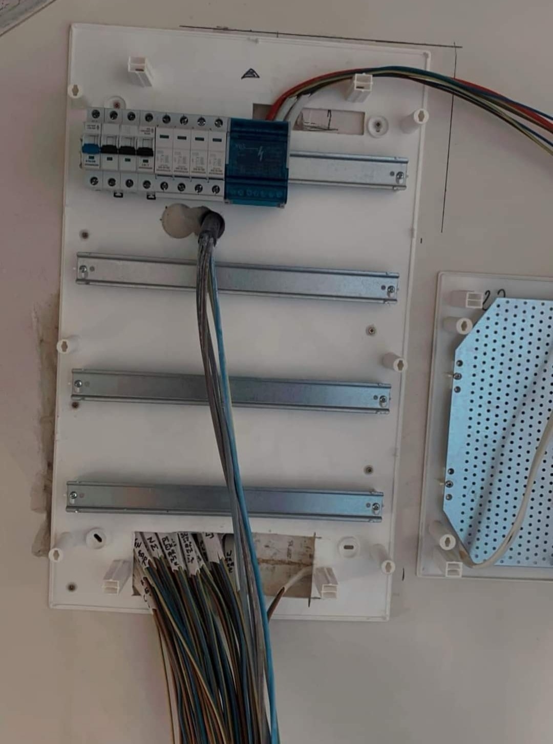 Otwarta, biała skrzynka rozdzielcza z zamontowanymi bezpiecznikami i listwami DIN, z wystającymi przewodami oznaczonymi opisami, widocznymi w dolnej części obudowy.