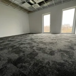 Nowo ułożona, ciemnoszara wykładzina dywanowa w płytkach w pustym biurze w Warszawie, widoczne okna z widokiem na drzewa i sufit podwieszany.