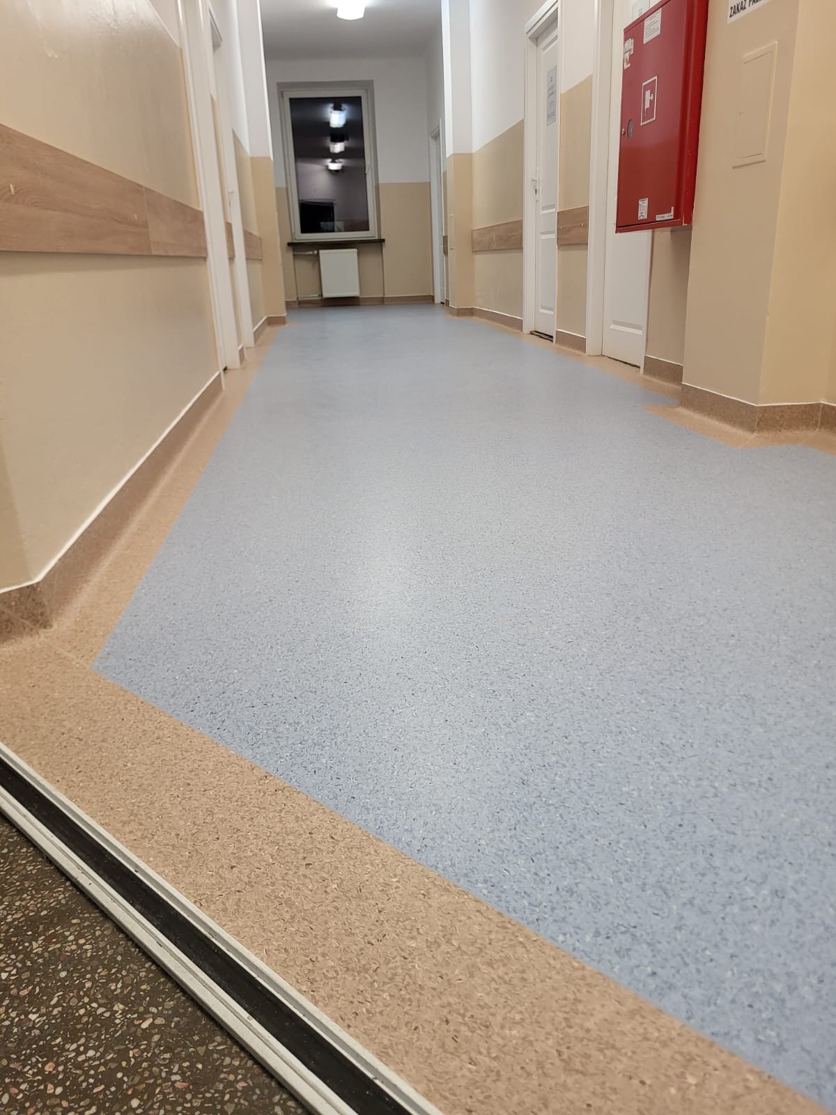 Jasnoniebieska wykładzina PCV w korytarzu placówki medycznej, z widocznymi listwami przypodłogowymi w kolorze beżowym i czerwonym hydrantem na ścianie.