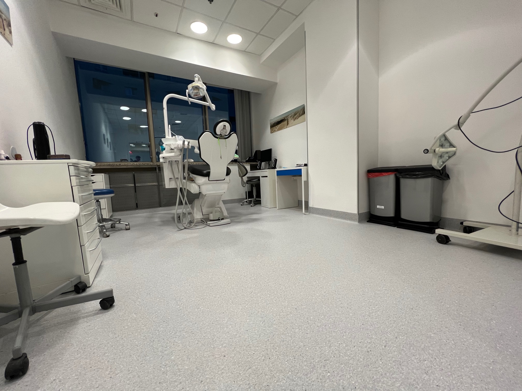 Jasnoszara, wzorzysta posadzka z PCV w gabinecie dentystycznym, widok na fotel dentystyczny i wyposażenie, duże okno w tle.
