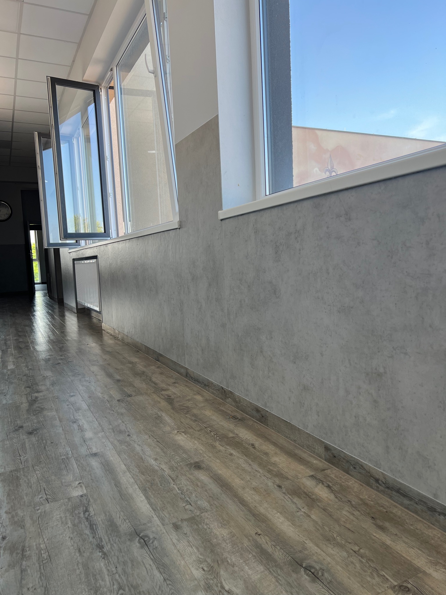 Korytarz z szarą podłogą imitującą drewno i szarymi ścianami, otwarte okna wpuszczają światło dzienne.