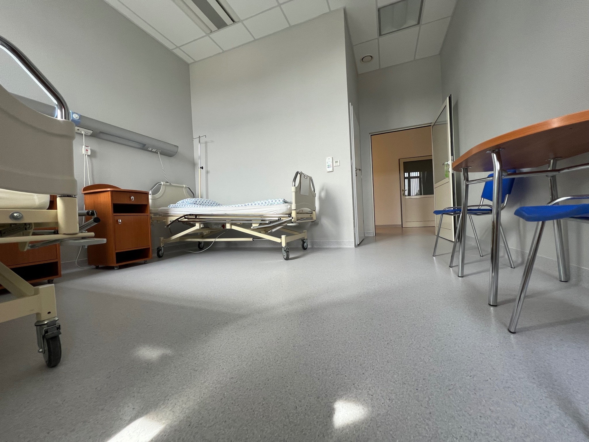 Szara, bezszwowa posadzka z drobnym wzorem w sterylnym pomieszczeniu szpitalnym, widoczny fragment łóżka medycznego, szafki nocnej i stołu z krzesłami.