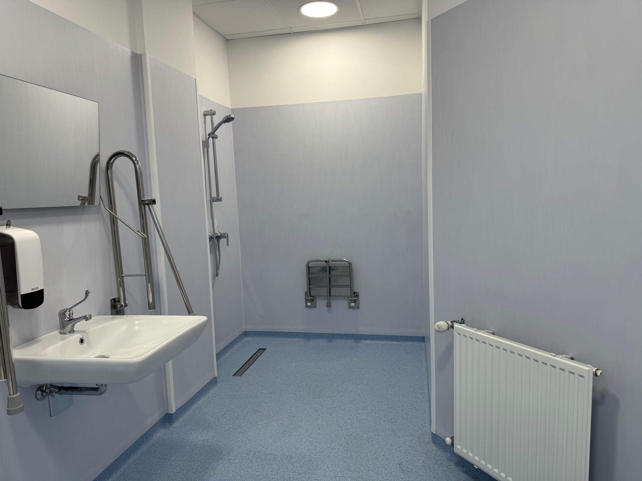 Łazienka przystosowana dla osób niepełnosprawnych z umywalką, poręczami, prysznicem ze składanym siedziskiem i niebieską wykładziną podłogową z odpływem liniowym w podłodze.