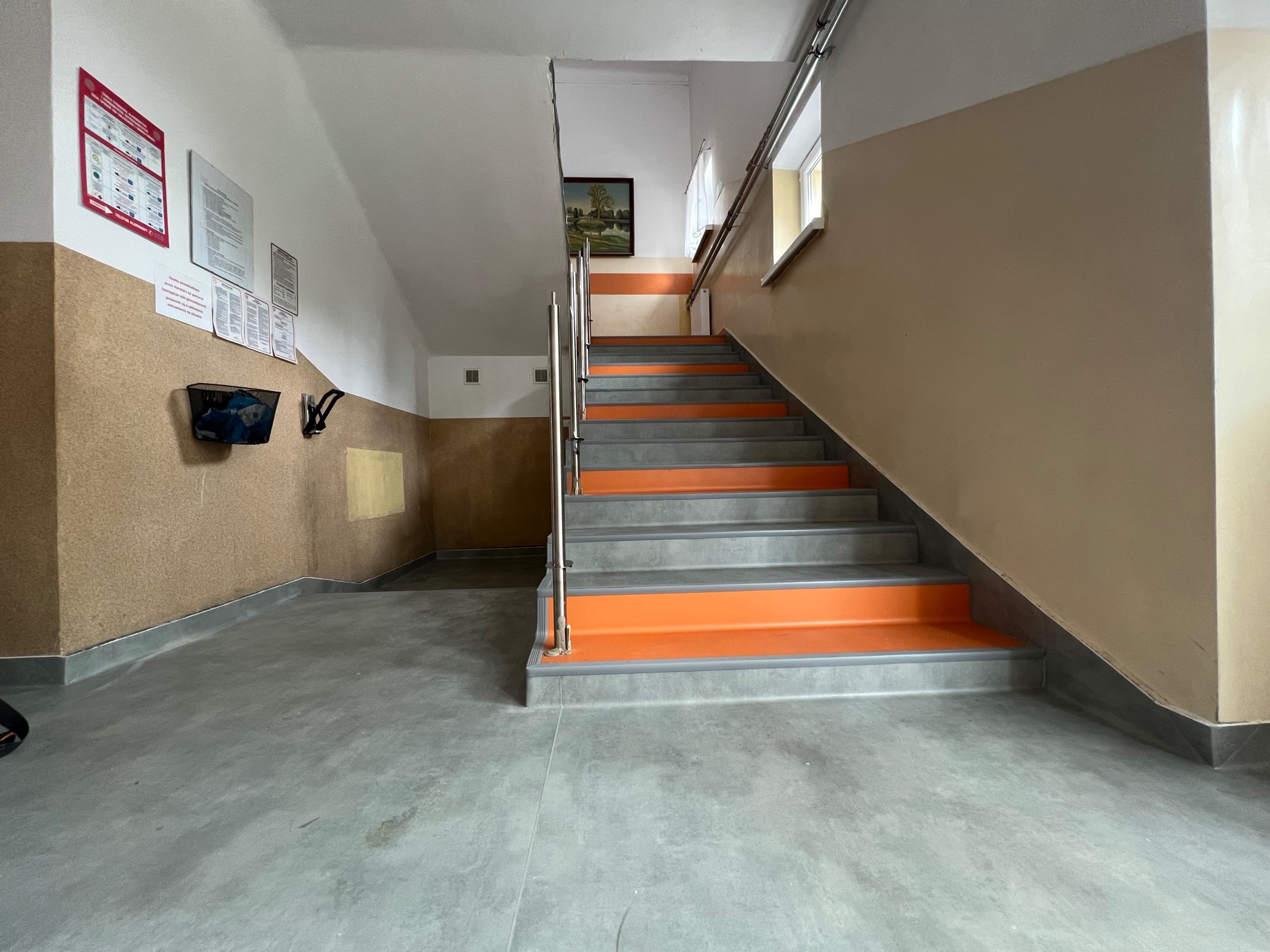 Klatka schodowa z szarymi schodami i pomarańczowymi podstopnicami, szara posadzka, ściany w kolorze beżowym i brązowym, na ścianie tablice informacyjne.