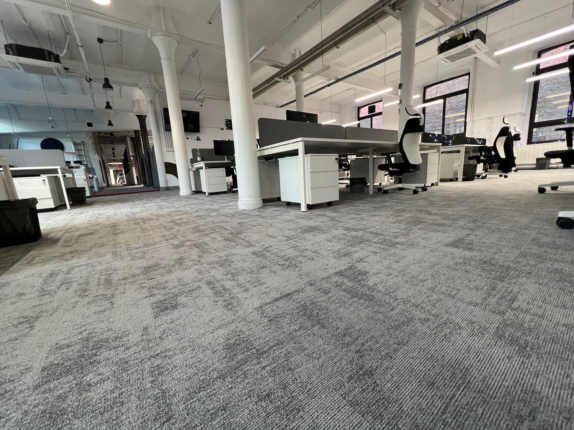 Biuro z szarą wykładziną dywanową w kwadraty, białe biurka z szufladami i kolumny wspierające sufit.